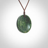 This piece is a medium sized oval round, cupped disc pendant. It was carved for us by Ric Moor from a lovely dark and varied green piece of New Zealand jade. It is suspended on a brown coloured braided cord that is length adjustable.